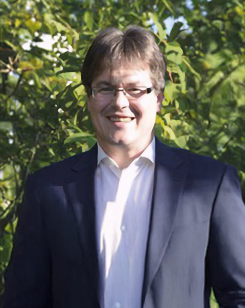 Ein Mann mit Brille, dunklem Anzug und weißem Hemd lächelt in die Kamera, während er im Freien vor grünem Laub steht.