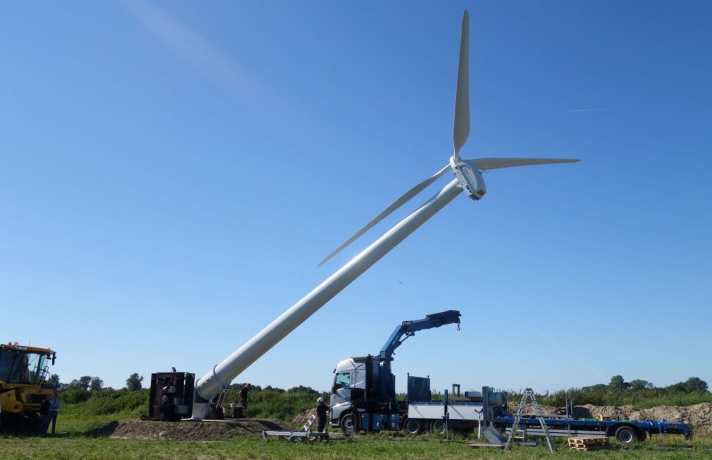 Eine große Windturbine wird auf einer Wiese zusammengebaut, Arbeiter und Fahrzeuge sind vor Ort und der Himmel ist blau.