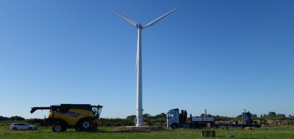 Eine Windkraftanlage steht auf einem Feld unter blauem Himmel, in der Nähe sind ein weißes Auto, ein gelber Mähdrescher und ein blauer Lkw geparkt.